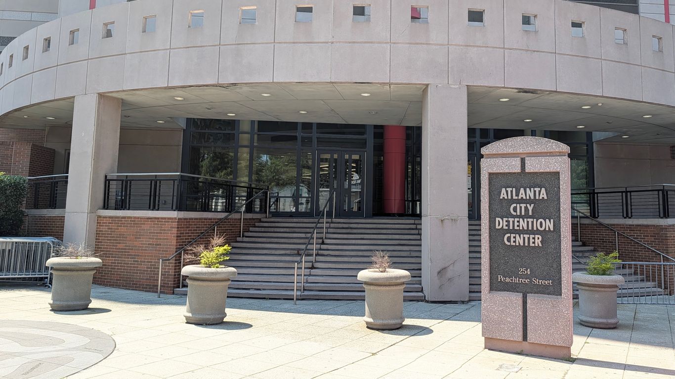 Exterior of the Atlanta City Detention Center: curved gray concrete facade with small square windows, glass doors, a red column, and stairs with railings; granite sign reads ATLANTA CITY DETENTION CENTER.
